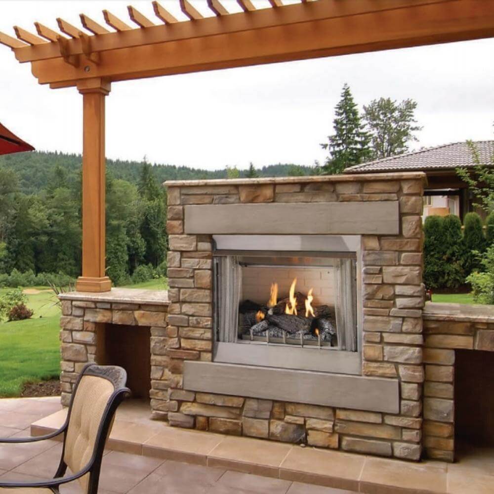 Empire Carol Rose Harmony Outdoor Stainless Steel Gas Log Insert - Outdoor stone fireplace with flames on a patio area with trees and grass in the background