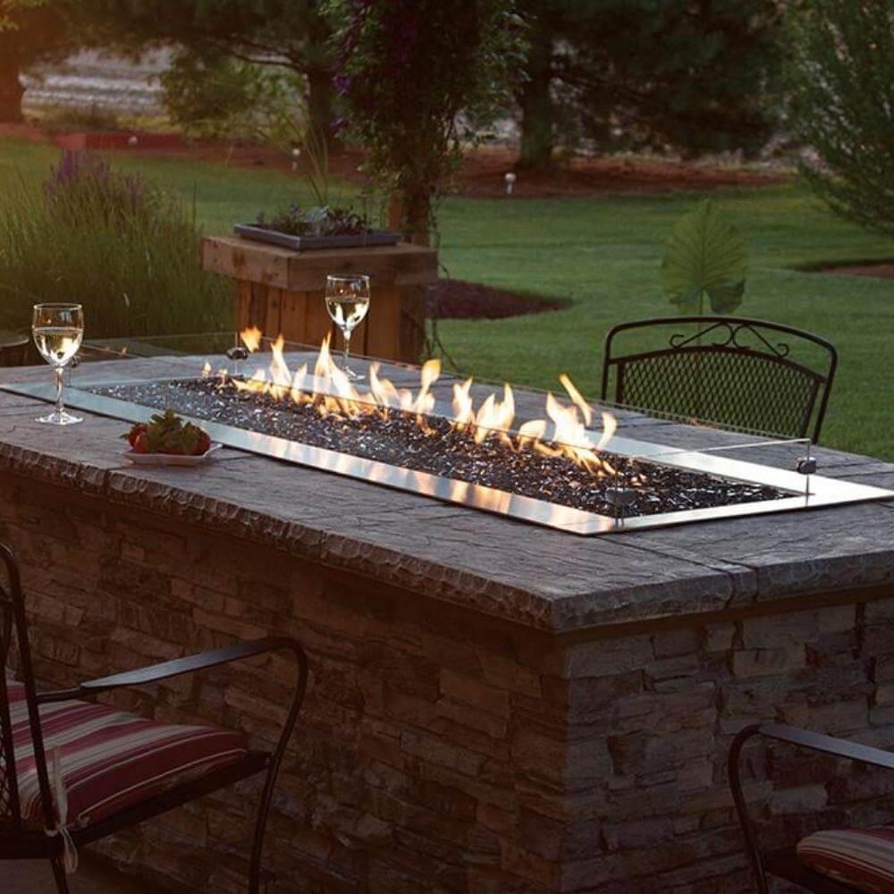 Empire Carol Rose Outdoor Stainless Steel Linear Gas Burner - Outdoor fire pit table with flames, glasses, and a garden view.