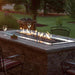 Empire Carol Rose Outdoor Stainless Steel Linear Gas Burner - Outdoor fire pit table with flames, glasses, and a garden view.