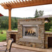 Empire Carol Rose Premium Outdoor Vent Free Gas Fireplace - Outdoor stone fireplace with flames on a patio area with trees and grass in the background