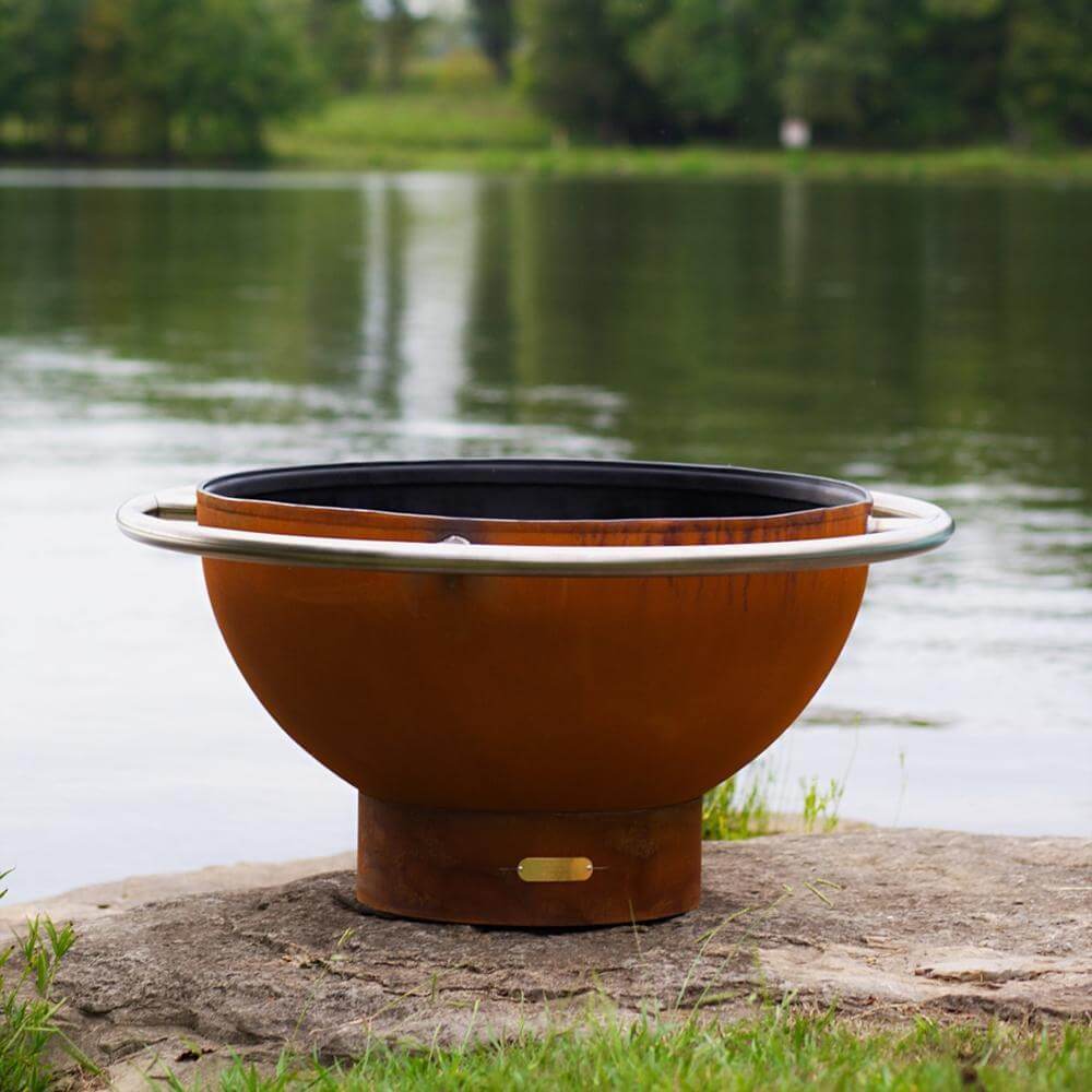 Fire Pit Art Bella Luna 48-Inch Handcrafted Carbon Steel Gas Fire Pit - Rust-colored fire pit on a rock by a lake with trees in the background