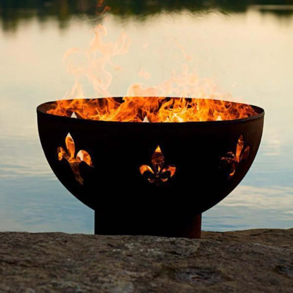 Fire Pit Art Fleur de Lis 36-Inch Handcrafted Carbon Steel Fire Pit (FDL) - Fire pit with flames burning in it, placed on a rocky surface by a body of water.