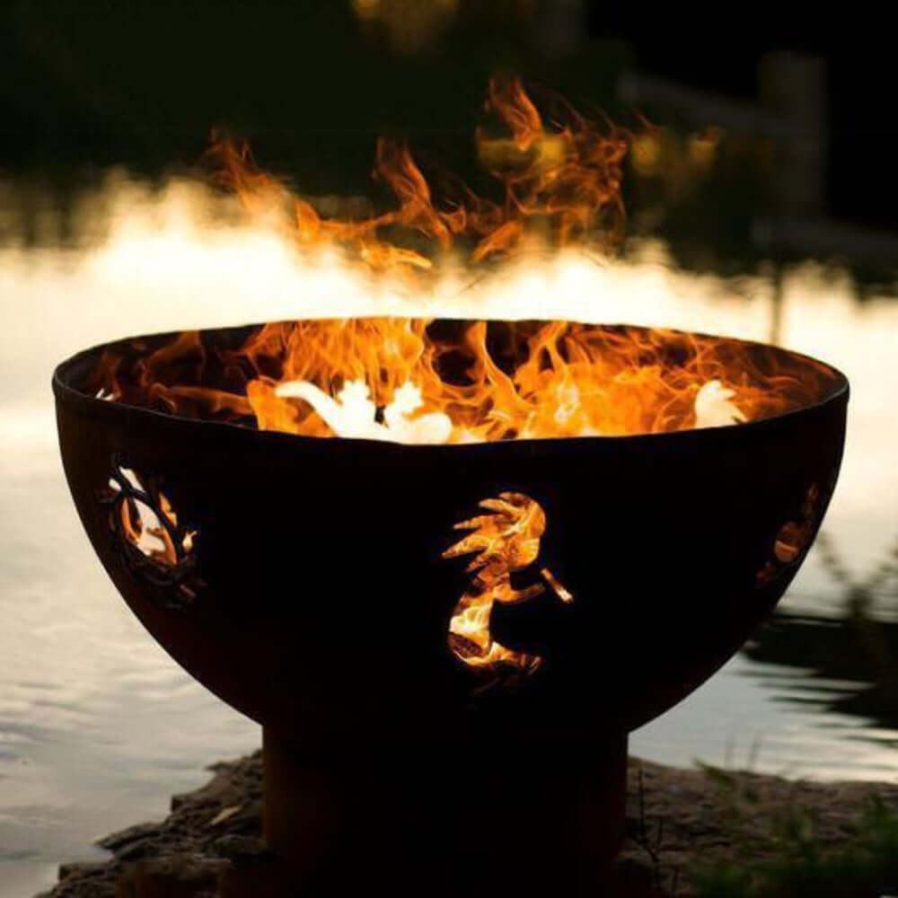 Fire Pit Art Kokopelli 36-Inch Handcrafted Carbon Steel Fire Pit (KO) - Fire pit with flames burning in it, placed by a body of water.