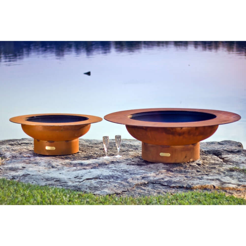 Fire Pit Art Magnum 54-Inch Handcrafted Carbon Steel Fire Pit (MAG) - Two copper-colored fire pits on a rocky shore with water in the background