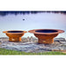 Fire Pit Art Magnum 54-Inch Handcrafted Carbon Steel Fire Pit (MAG) - Two copper-colored fire pits on a rocky shore with water in the background