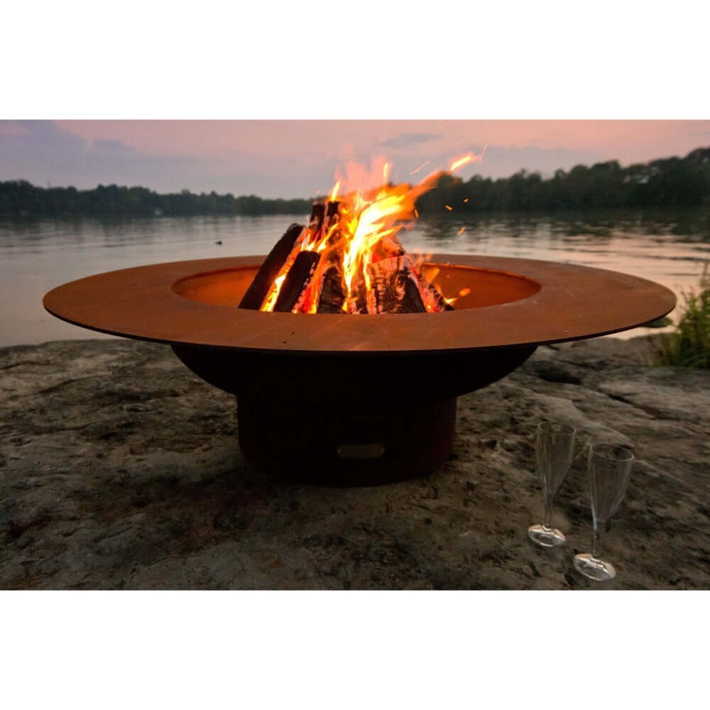 Fire Pit Art Magnum 54-Inch Handcrafted Carbon Steel Fire Pit (MAG) - Fire pit with flames burning on a rock by a body of water