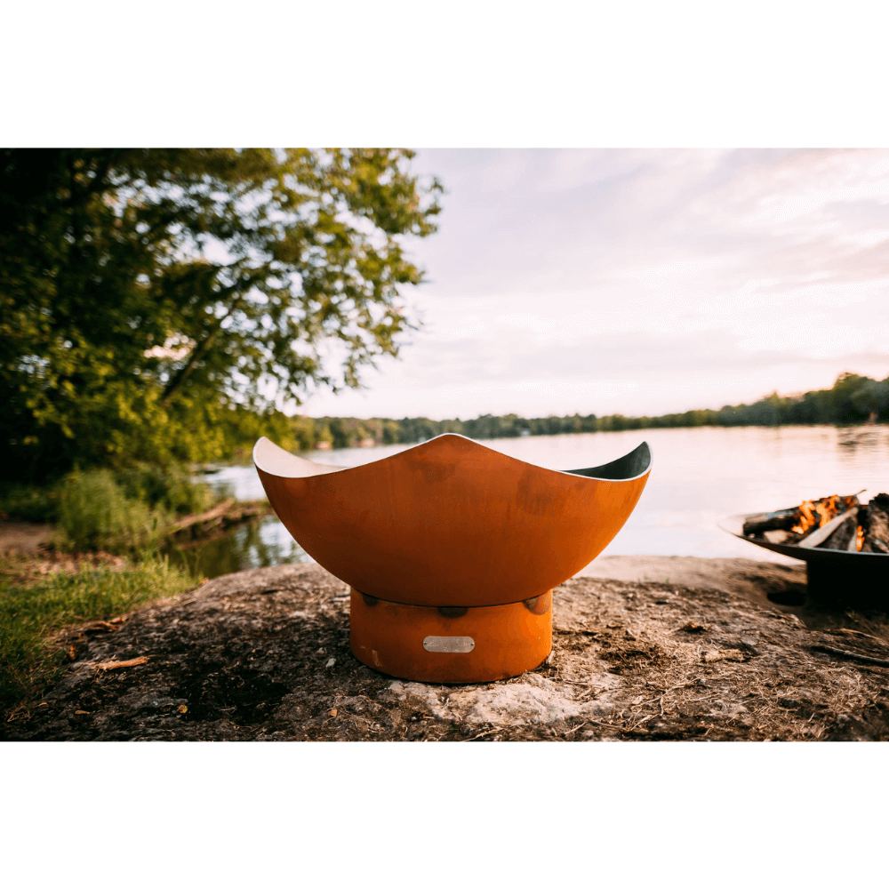 Fire Pit Art Manta Ray 36-Inch Handcrafted Carbon Steel Gas Fire Pit - Orange fire pit on a rock by a lake with trees in the background
