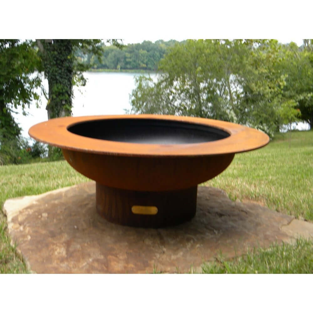 Fire Pit Art Saturn 40-Inch Handcrafted Carbon Steel Gas Fire Pit - Rust-colored fire pit on a stone platform with a lake and trees in the background