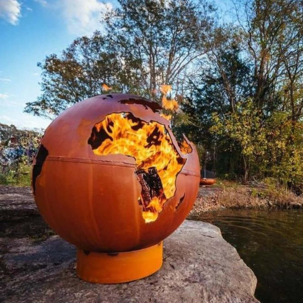 Fire Pit Art Third Rock Globe Shaped 36-Inch Carbon Steel Fire Pit (TR) - Spherical fire pit with flames burning outdoors near a body of water and trees.