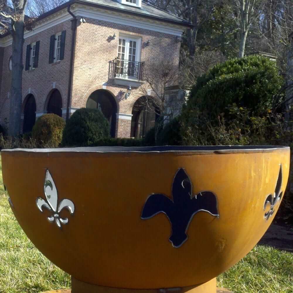 Fire Pit Art Fleur de Lis 36-Inch Handcrafted Carbon Steel Fire Pit (FDL) - Yellow fire pit with fleur-de-lis designs in front of a brick house.