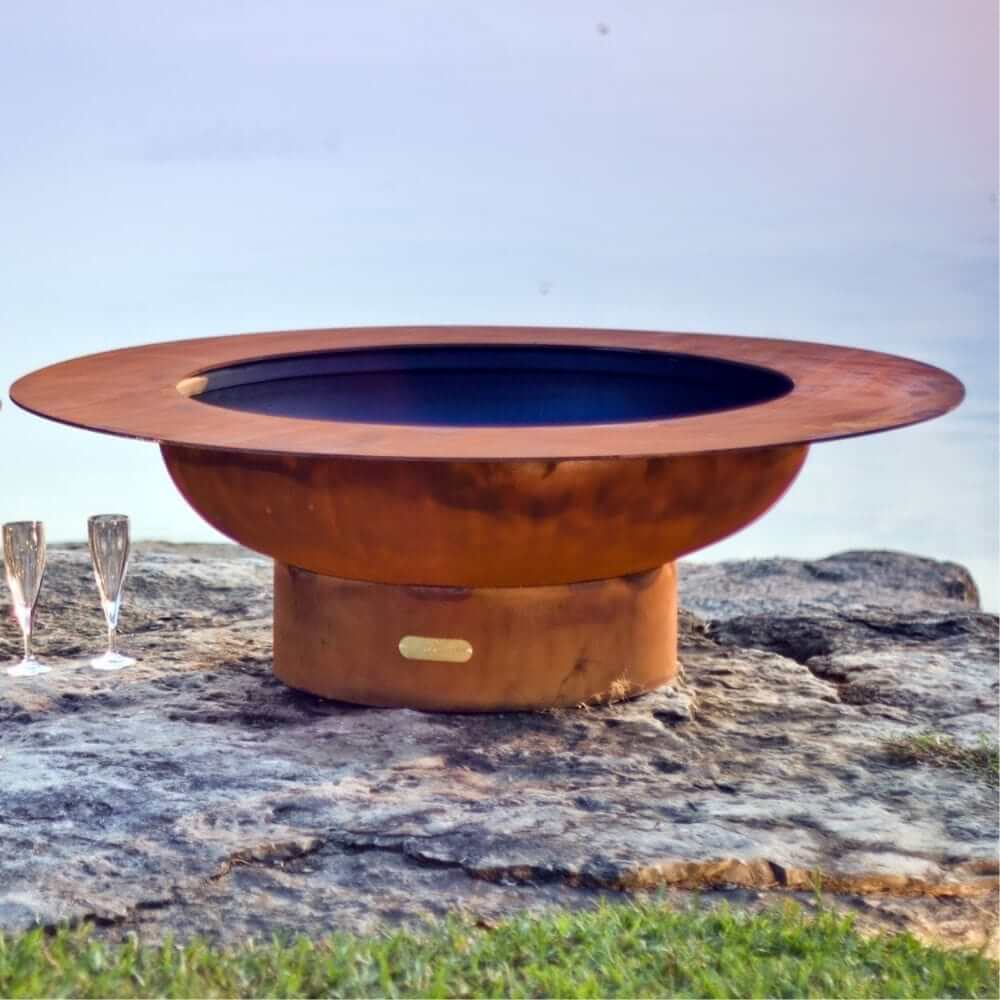 Fire Pit Art Magnum 54-Inch Handcrafted Carbon Steel Gas Fire Pit - Copper fire pit on a rocky surface with two glasses nearby