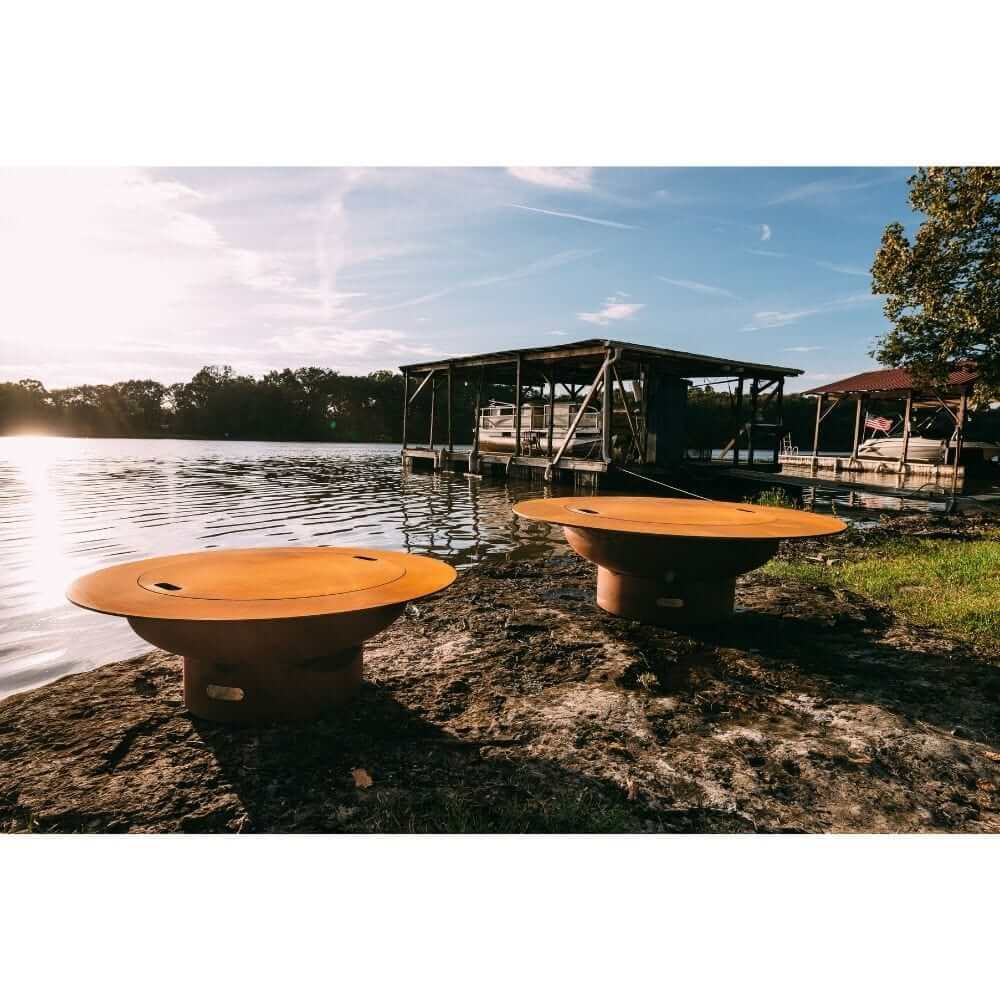 Fire Pit Art Magnum 54-Inch Handcrafted Carbon Steel Gas Fire Pit - Two round metal fire pits on a lakeside dock with a scenic background.