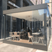 Modern glass-walled dining space with people seated at tables in an outdoor setting.