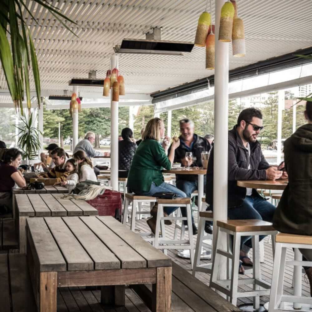 Outdoor dining area with people enjoying food and drinks under electric heaters.